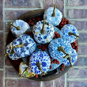 Puede incluir: Una colecci&oacute;n de nueve calabazas blancas decoradas con motivos florales azules y dorados. Las calabazas est&aacute;n dispuestas en una bandeja de madera con bayas rojas esparcidas a su alrededor.