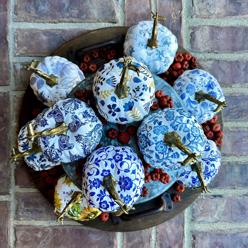 Puede incluir: Una colecci&oacute;n de nueve calabazas blancas decoradas con motivos florales azules y dorados. Las calabazas est&aacute;n dispuestas en una bandeja de madera con bayas rojas esparcidas a su alrededor.
