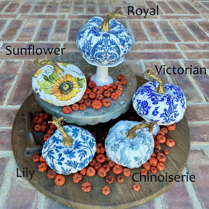 Puede incluir: Cuatro calabazas blancas decoradas con motivos florales azules y blancos. Las calabazas est&aacute;n dispuestas en un soporte de madera de varios niveles con capullos de flores naranjas secas. Las calabazas est&aacute;n etiquetadas como "Sunflower", "Royal", "Victorian" y "Chinoiserie".