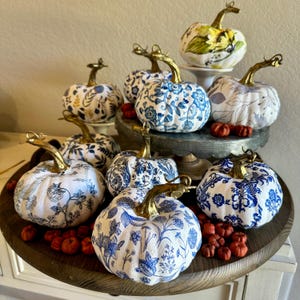 Puede incluir: Una colecci&oacute;n de calabazas con patrones azules y blancos con detalles dorados. Las calabazas est&aacute;n dispuestas en una bandeja de madera con bayas rojas esparcidas a su alrededor.