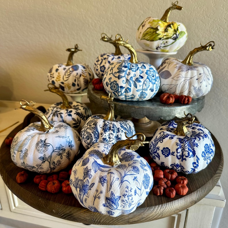 Puede incluir: Una colecci&oacute;n de calabazas con patrones azules y blancos con detalles dorados. Las calabazas est&aacute;n dispuestas en una bandeja de madera con bayas rojas esparcidas a su alrededor.