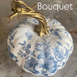 Peut inclure: Citrouille d&eacute;corative avec une base blanche et un motif floral bleu. La tige est dor&eacute;e et pr&eacute;sente un design incurv&eacute;. Le mot "Bouquet" est imprim&eacute; en blanc en haut &agrave; droite de la citrouille.