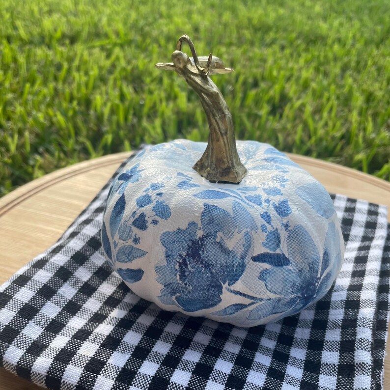 Peut inclure: Une citrouille d&eacute;corative avec un motif floral bleu sur fond blanc. La citrouille a une tige dor&eacute;e et repose sur un tissu &agrave; carreaux noir et blanc. C'est un article de d&eacute;coration saisonnier.