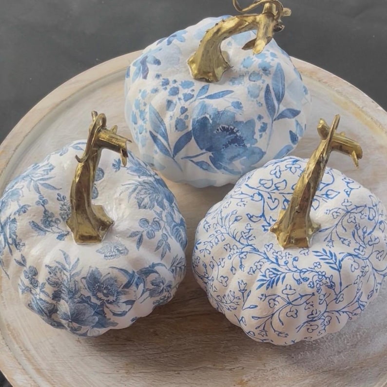 Peut inclure: Trois citrouilles d&eacute;coratives avec des motifs floraux blancs et bleus. Chaque citrouille a une tige dor&eacute;e. Les citrouilles sont dispos&eacute;es sur un plateau en bois clair, cr&eacute;ant une d&eacute;coration d'automne.