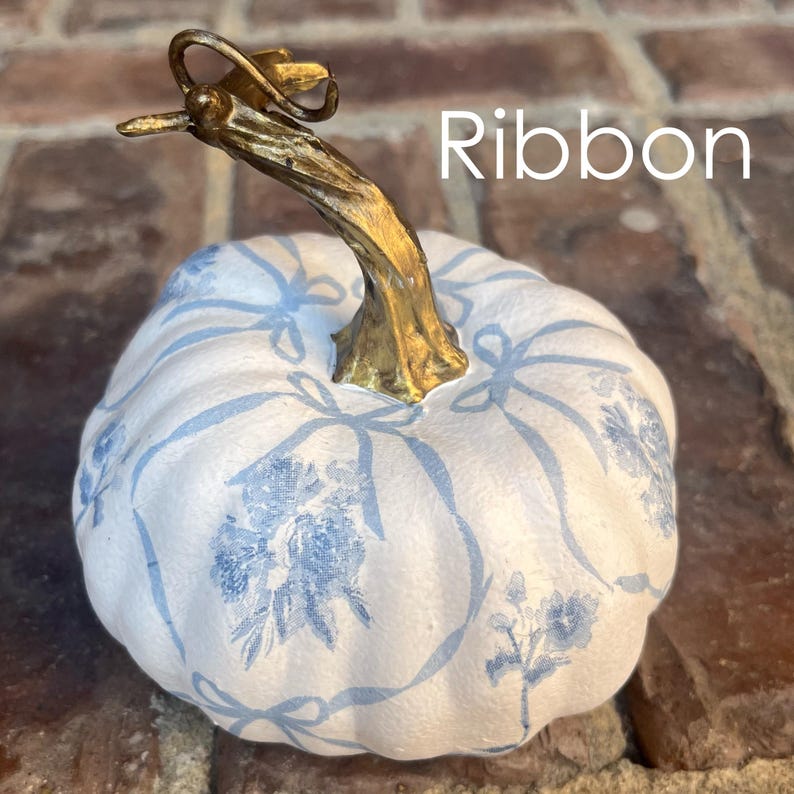 Peut inclure: Une citrouille blanche d&eacute;cor&eacute;e de motifs floraux et de rubans bleus. Elle pr&eacute;sente une tige dor&eacute;e et le mot "Ribbon". Cet article de saison est id&eacute;al pour la d&eacute;coration automnale, ajoutant une touche d'&eacute;l&eacute;gance.