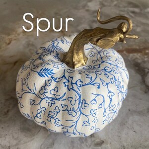 Peut inclure: Une citrouille d&eacute;corative avec une base blanche et un motif floral bleu. La tige est de couleur or, et le mot "Spur" est &eacute;crit en lettres blanches en haut &agrave; gauche de la citrouille. Cet article sur le th&egrave;me de l'automne est parfait pour la d&eacute;coration int&eacute;rieure.