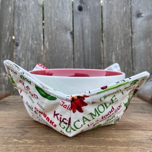 May include: White bowl cozy with a red rim, made of fabric with a white background and green and red text and images. The text includes "Guacamole", "Jala", "Cilantro", "Salt & Pepper", "Kicks", and "In a food processor".