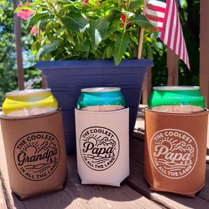 Puede incluir: Tres aislantes cil&iacute;ndricos para bebidas hechos de piel sint&eacute;tica en color tostado, blanco y marr&oacute;n. Cada uno presenta un gr&aacute;fico circular con el texto "The Coolest Grandpa in All the Land" o "The Coolest Papa in All the Land" en tinta negra.
