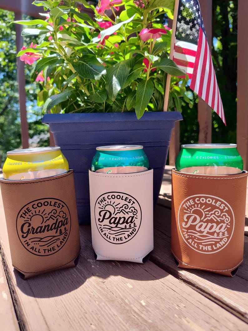 Puede incluir: Tres aislantes cil&iacute;ndricos para bebidas hechos de piel sint&eacute;tica en color tostado, blanco y marr&oacute;n. Cada uno presenta un gr&aacute;fico circular con el texto "The Coolest Grandpa in All the Land" o "The Coolest Papa in All the Land" en tinta negra.