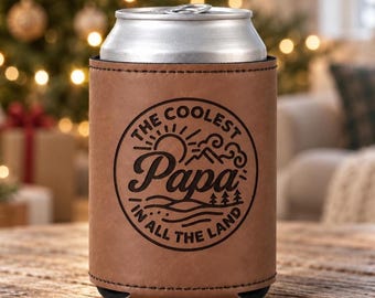 May include: A brown leatherette can cooler sleeve featuring a black circular graphic with a sun, mountains, and trees. The text reads THE COOLEST Papa IN ALL THE LAND. The sleeve is wrapped around a silver aluminum beverage can.