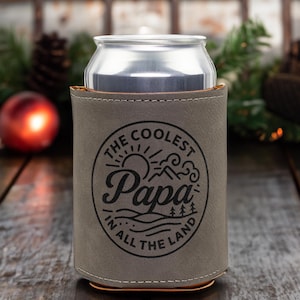 Puede incluir: Una funda enfriadora de latas de piel sint&eacute;tica gris con un gr&aacute;fico circular negro que muestra un sol, monta&ntilde;as y &aacute;rboles. El texto dice THE COOLEST Papa IN ALL THE LAND. La funda envuelve una lata de bebida de aluminio plateado con costuras.