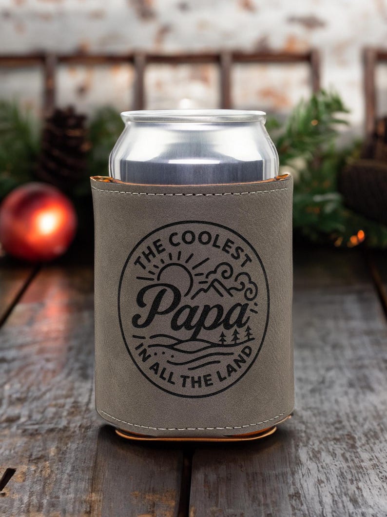 Puede incluir: Una funda enfriadora de latas de piel sint&eacute;tica gris con un gr&aacute;fico circular negro que muestra un sol, monta&ntilde;as y &aacute;rboles. El texto dice THE COOLEST Papa IN ALL THE LAND. La funda envuelve una lata de bebida de aluminio plateado con costuras.