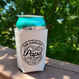 Puede incluir: Un enfriador de latas blanco con un gr&aacute;fico circular negro que incluye el texto "THE COOLEST Papa IN ALL THE LAND" rodeado de ilustraciones de un sol, monta&ntilde;as y olas. El enfriador sostiene una lata de aluminio de color verde azulado.