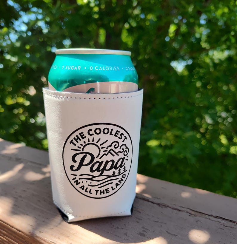 Puede incluir: Un enfriador de latas blanco con un gr&aacute;fico circular negro que incluye el texto "THE COOLEST Papa IN ALL THE LAND" rodeado de ilustraciones de un sol, monta&ntilde;as y olas. El enfriador sostiene una lata de aluminio de color verde azulado.