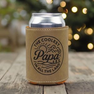Puede incluir: Una funda de cuero color canela para latas con un gr&aacute;fico circular negro que incluye el texto "THE COOLEST Papa IN ALL THE LAND" rodeando una ilustraci&oacute;n de un sol, monta&ntilde;as y pinos, conteniendo una lata de bebida de aluminio plateado.