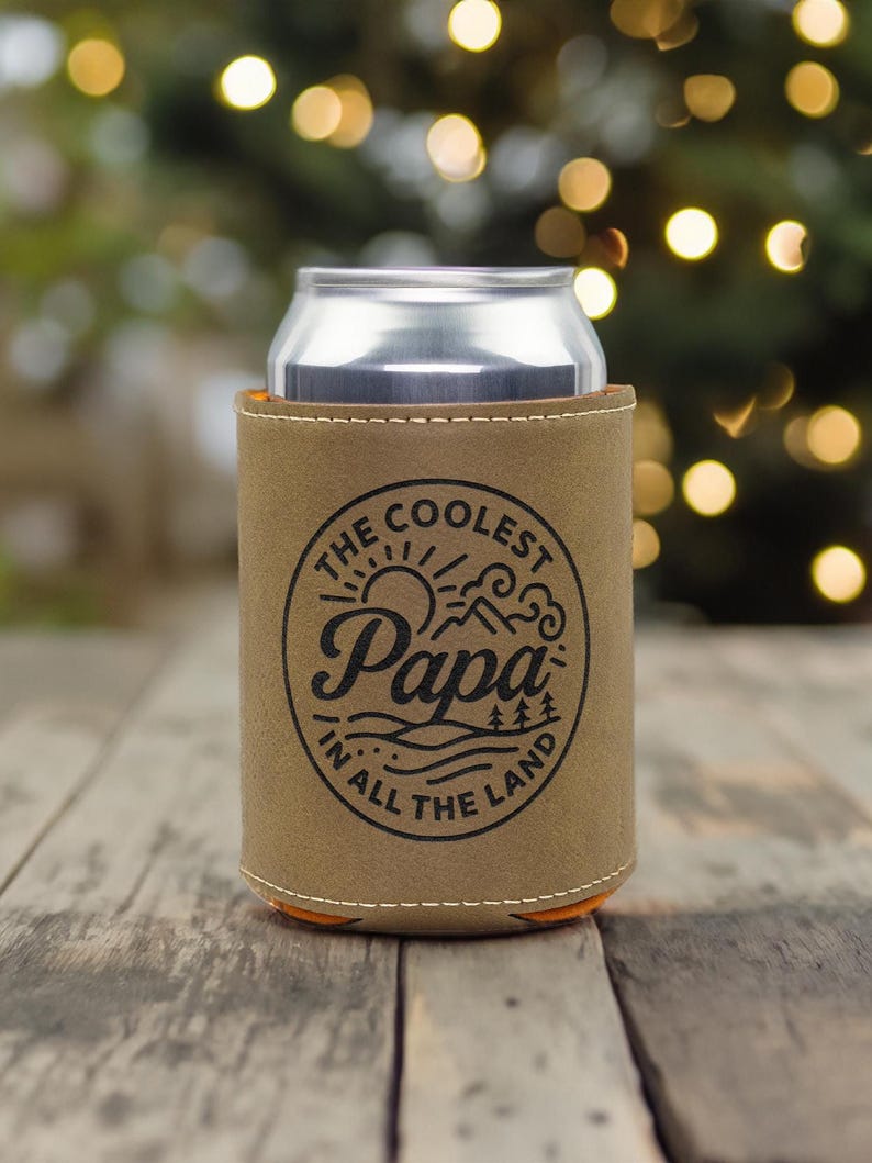 Puede incluir: Una funda de cuero color canela para latas con un gr&aacute;fico circular negro que incluye el texto "THE COOLEST Papa IN ALL THE LAND" rodeando una ilustraci&oacute;n de un sol, monta&ntilde;as y pinos, conteniendo una lata de bebida de aluminio plateado.