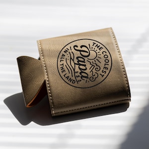 Puede incluir: Un enfriador de latas de piel sint&eacute;tica color canela con un gr&aacute;fico circular negro que incluye el texto "THE COOLEST PAPA IN ALL THE LAND" junto a ilustraciones de un sol, monta&ntilde;as, olas y &aacute;rboles. La funda tiene costuras visibles.