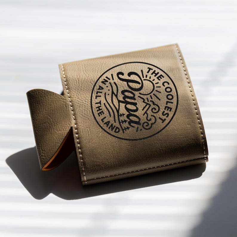 Puede incluir: Un enfriador de latas de piel sint&eacute;tica color canela con un gr&aacute;fico circular negro que incluye el texto "THE COOLEST PAPA IN ALL THE LAND" junto a ilustraciones de un sol, monta&ntilde;as, olas y &aacute;rboles. La funda tiene costuras visibles.