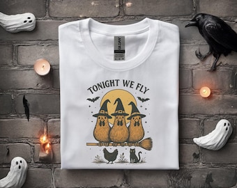 May include: White t-shirt featuring a Halloween-themed graphic. The design includes three cartoon chickens wearing witch hats, riding a broomstick, with the text "TONIGHT WE FLY". A black cat and a chicken are also depicted.