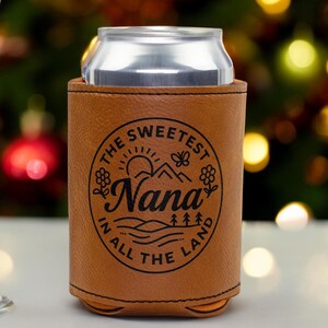 Puede incluir: Una funda enfriadora de latas de piel sint&eacute;tica color canela con un gr&aacute;fico circular negro que incluye el texto "THE SWEETEST Nana IN ALL THE LAND" rodeando una ilustraci&oacute;n de sol, monta&ntilde;as, flores, una mariposa y &aacute;rboles, con una lata de aluminio.