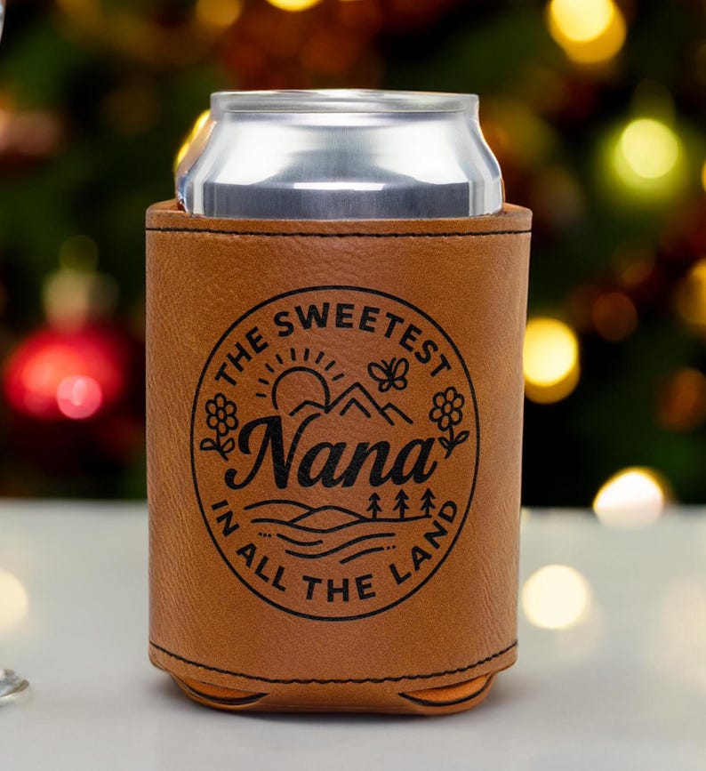 Puede incluir: Una funda enfriadora de latas de piel sint&eacute;tica color canela con un gr&aacute;fico circular negro que incluye el texto "THE SWEETEST Nana IN ALL THE LAND" rodeando una ilustraci&oacute;n de sol, monta&ntilde;as, flores, una mariposa y &aacute;rboles, con una lata de aluminio.