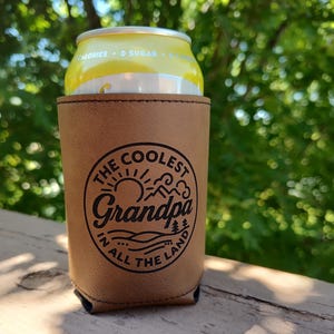 Puede incluir: Una funda de cuero color canela para latas con un gr&aacute;fico circular negro que muestra un sol, monta&ntilde;as y &aacute;rboles. El texto dice THE COOLEST Grandpa IN ALL THE LAND. La funda sostiene una lata de bebida amarilla con el texto 0 SUGAR.