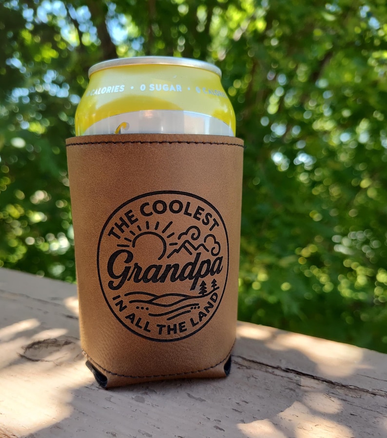 Puede incluir: Una funda de cuero color canela para latas con un gr&aacute;fico circular negro que muestra un sol, monta&ntilde;as y &aacute;rboles. El texto dice THE COOLEST Grandpa IN ALL THE LAND. La funda sostiene una lata de bebida amarilla con el texto 0 SUGAR.