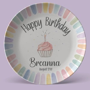 Könnte beinhalten: Ein weißer Keramikteller mit einem bunten Rand aus pastellfarbenen Pinselstrichen im Aquarellstil. In der Mitte befindet sich ein rosa Cupcake mit einer brennenden Kerze, der Text Happy Birthday, der Name Breanna und das Datum August 21st.