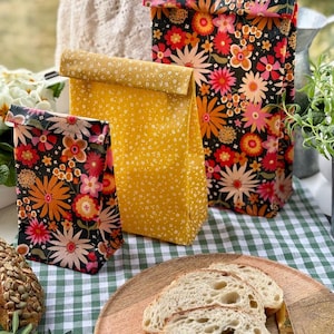 May include: Three reusable lunch bags are displayed. One is yellow with small white flowers, and the other two are black with a vibrant floral design. Sliced bread on a wooden plate and a knife are in the foreground.