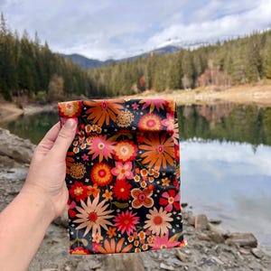 Pode incluir: Uma bolsa preta com um padr&atilde;o floral vibrante em tons de laranja, rosa e vermelho. A bolsa &eacute; segurada contra um cen&aacute;rio de um lago e montanhas.