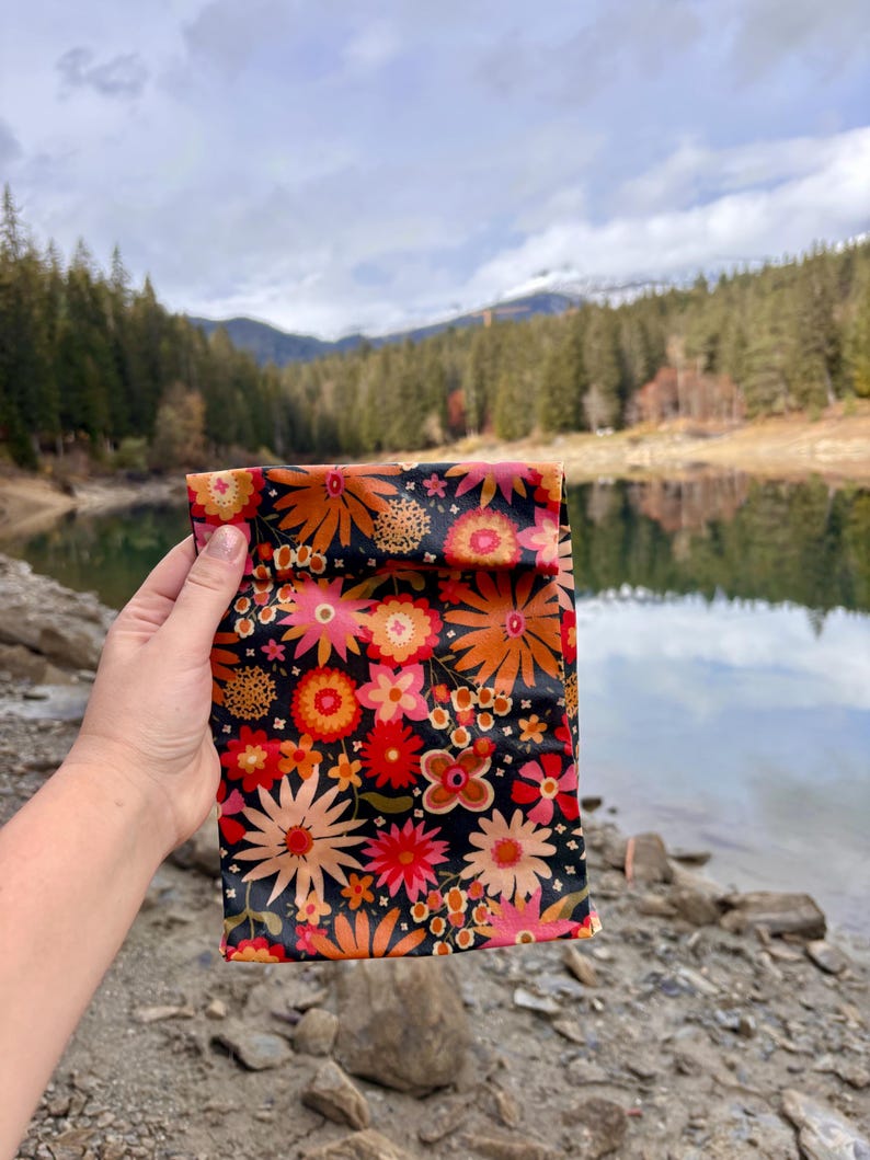 Pode incluir: Uma bolsa preta com um padr&atilde;o floral vibrante em tons de laranja, rosa e vermelho. A bolsa &eacute; segurada contra um cen&aacute;rio de um lago e montanhas.