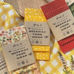 May include: A collection of reusable beeswax food wraps in various patterns including yellow floral, red with white dots, green with white dots, and a multi-colored botanical print. Each wrap features a brown paper band labeled Wrap it.