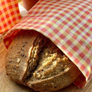 Pode incluir: Um p&atilde;o rec&eacute;m-assado, parcialmente embrulhado em um pano xadrez vermelho e amarelo. O p&atilde;o tem uma crosta marrom escura e sementes vis&iacute;veis. Um vaso de terracota com folhas verdes est&aacute; ao fundo.