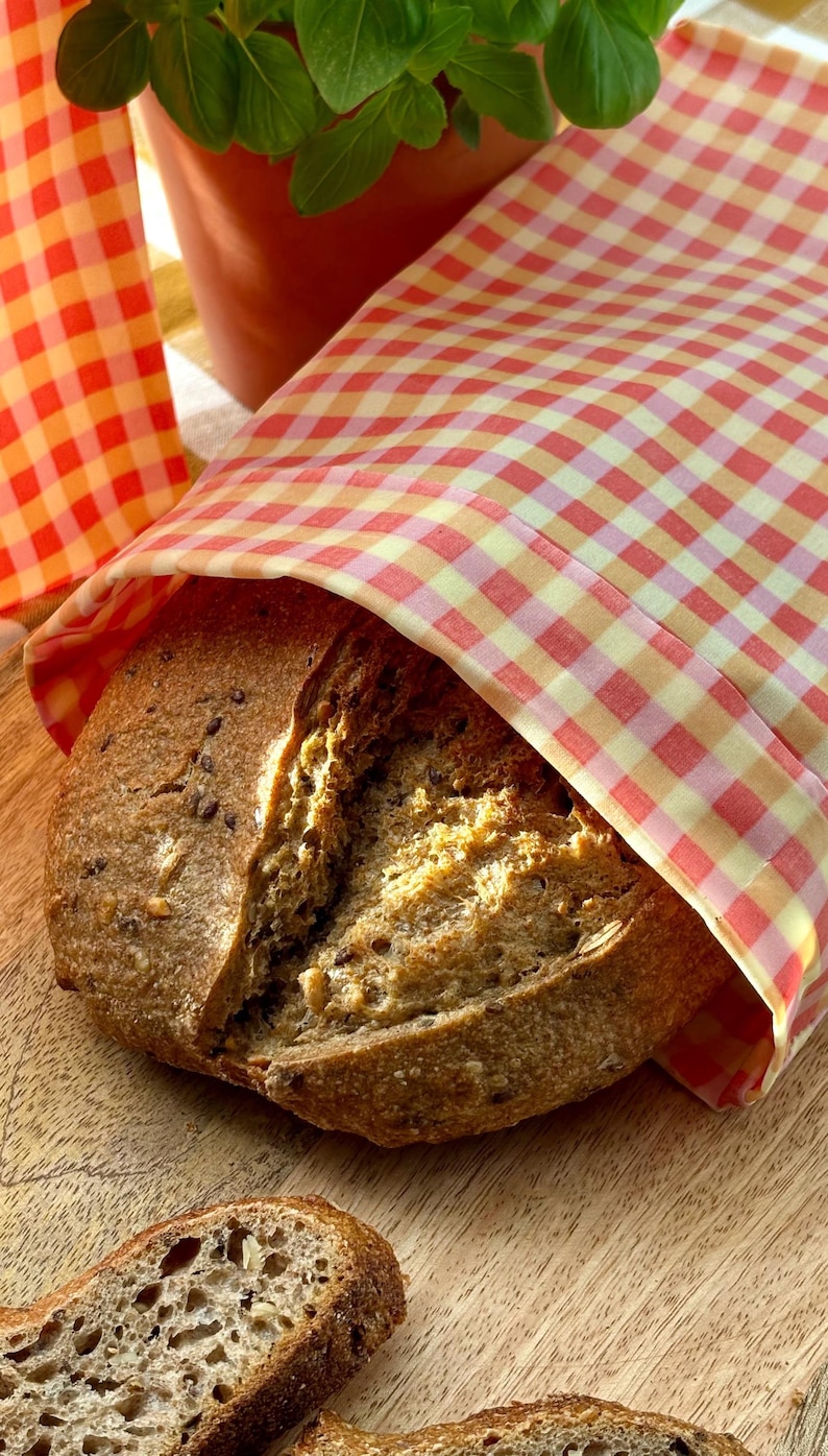 Pode incluir: Um p&atilde;o rec&eacute;m-assado, parcialmente embrulhado em um pano xadrez vermelho e amarelo. O p&atilde;o tem uma crosta marrom escura e sementes vis&iacute;veis. Um vaso de terracota com folhas verdes est&aacute; ao fundo.