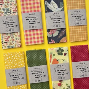 May include: Eight rectangular beeswax food wraps with various patterns including floral, gingham, bird, and vegetable prints. Each wrap features a paper band labelled Wrap it Natural Alternative Reusable Beeswax Food Wrap with bee icons.