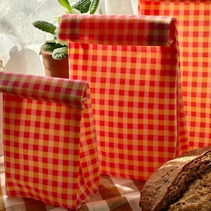 Pode incluir: S&atilde;o exibidas tr&ecirc;s sacolas de almo&ccedil;o xadrez vermelhas e laranja de tamanhos variados. As sacolas s&atilde;o dobradas na parte superior. Um p&atilde;o est&aacute; sobre uma toalha de mesa xadrez marrom e branca. Uma planta em vaso est&aacute; ao fundo.