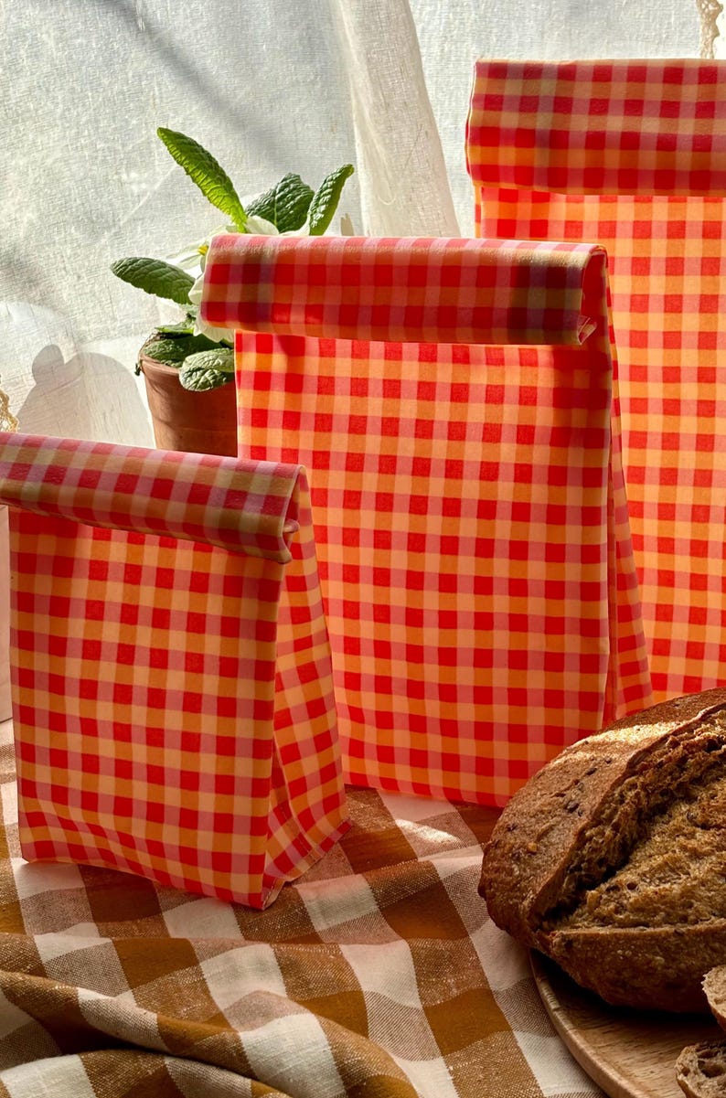 Pode incluir: S&atilde;o exibidas tr&ecirc;s sacolas de almo&ccedil;o xadrez vermelhas e laranja de tamanhos variados. As sacolas s&atilde;o dobradas na parte superior. Um p&atilde;o est&aacute; sobre uma toalha de mesa xadrez marrom e branca. Uma planta em vaso est&aacute; ao fundo.