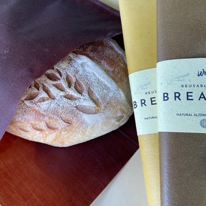 Pu&ograve; includere: Una pagnotta di pane artigianale con un disegno a foglie &egrave; parzialmente visibile all'interno di un sacchetto per il pane bordeaux scuro. Due involucri per il pane riutilizzabili sono accanto al pane, uno giallo e uno marrone, con etichette che recitano "REUSABLE BREAD WRAP".