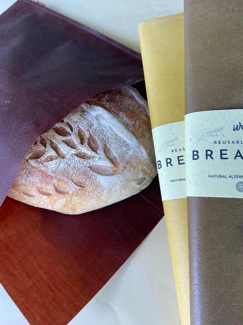 Pu&ograve; includere: Una pagnotta di pane artigianale con un disegno a foglie &egrave; parzialmente visibile all'interno di un sacchetto per il pane bordeaux scuro. Due involucri per il pane riutilizzabili sono accanto al pane, uno giallo e uno marrone, con etichette che recitano "REUSABLE BREAD WRAP".