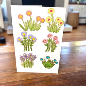 May include: A white greeting card featuring colorful hand-drawn illustrations of various flowers. The flowers include yellow, pink, purple, and blue blooms with green stems and leaves. The card is displayed on a wooden surface.