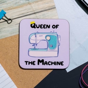 May include: A purple square coaster with a white outline and the text "QUEEN OF THE MACHINE" in black. A cartoon drawing of a sewing machine with a yellow crown above the text.