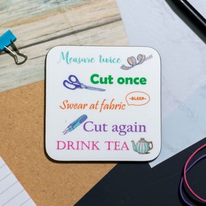 May include: White square coaster with the text "Measure twice, Cut once, Swear at fabric -BLEEP-, Cut again, DRINK TEA". The coaster features illustrations of scissors, a measuring tape, and a teapot.