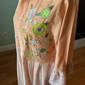 May include: Peach-colored button-down shirt dress with floral applique details. The dress features long sleeves with plaid cuffs and a patterned skirt. The floral appliques are in shades of green, orange, and blue.