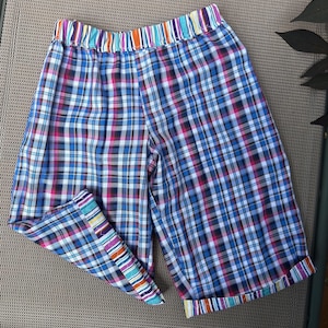 May include: Colorful plaid shorts with a striped waistband and cuffs. The shorts feature a blue, pink, and white plaid pattern. The waistband and cuffs have a vibrant striped design with various colors. These shorts are a casual summer garment.