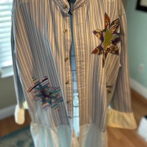 以下が含まれることがあります： マンダリンカラーの青いストライプのロングスリーブシャツ。シャツの前身頃には、カラフルな布のアップリケが2つ付いています。