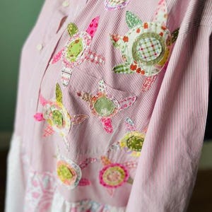 May include: A pink and white striped shirt with a white lace trim. The shirt is decorated with colorful fabric flowers.