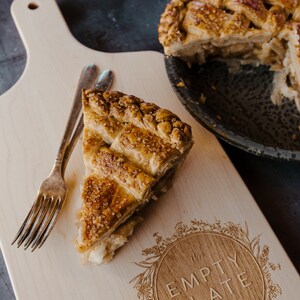 May include: A wooden cutting board with a slice of apple pie on it. The board is engraved with the words "The Empty Plate Est 2020" in a circle surrounded by floral designs. There are two forks on the board.