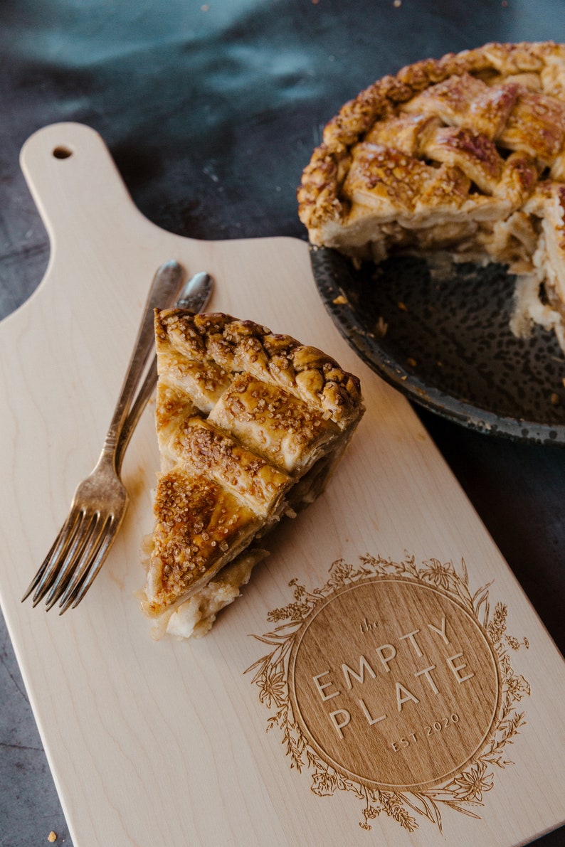May include: A wooden cutting board with a slice of apple pie on it. The board is engraved with the words "The Empty Plate Est 2020" in a circle surrounded by floral designs. There are two forks on the board.