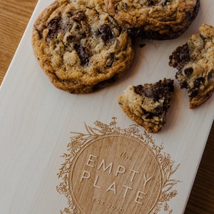 May include: A wooden cutting board with the text "The Empty Plate Est 2020" engraved in a circle surrounded by a floral design. There are four chocolate chip cookies on the board, one of which is broken in half.