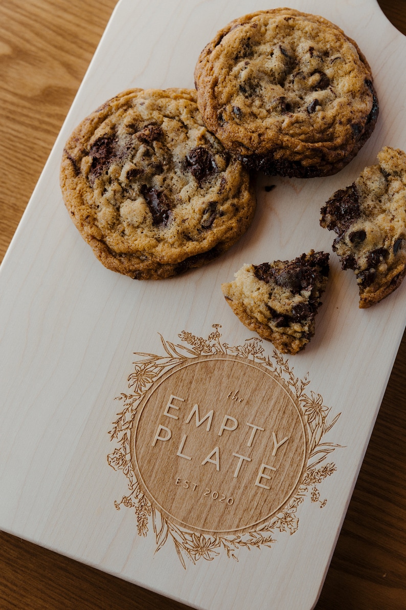 May include: A wooden cutting board with the text "The Empty Plate Est 2020" engraved in a circle surrounded by a floral design. There are four chocolate chip cookies on the board, one of which is broken in half.